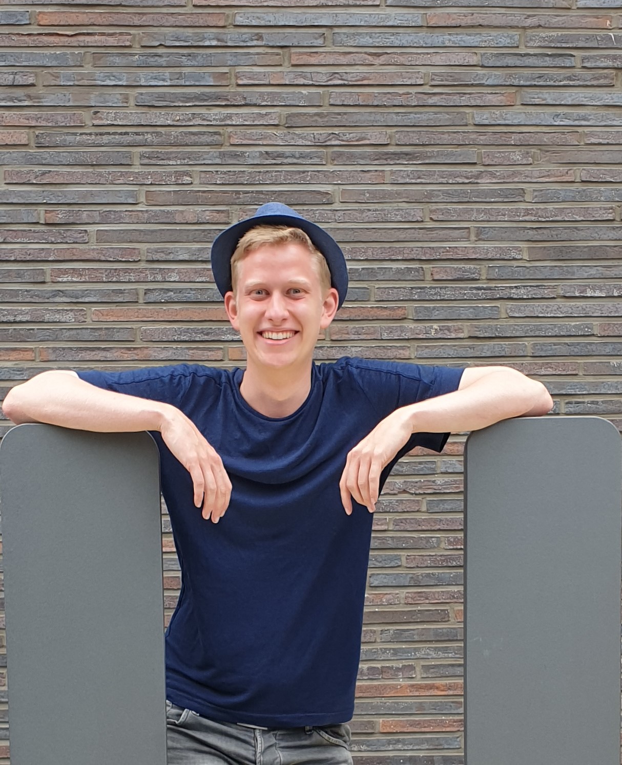 Testimonial student Henrik Remmert, young, male-appearing person with a blue T-shirt and hat, stands in front of a brick wall, supports his arms on the gray ends of the wall that protrude into the picture on the left and right, the person smiles into the camera